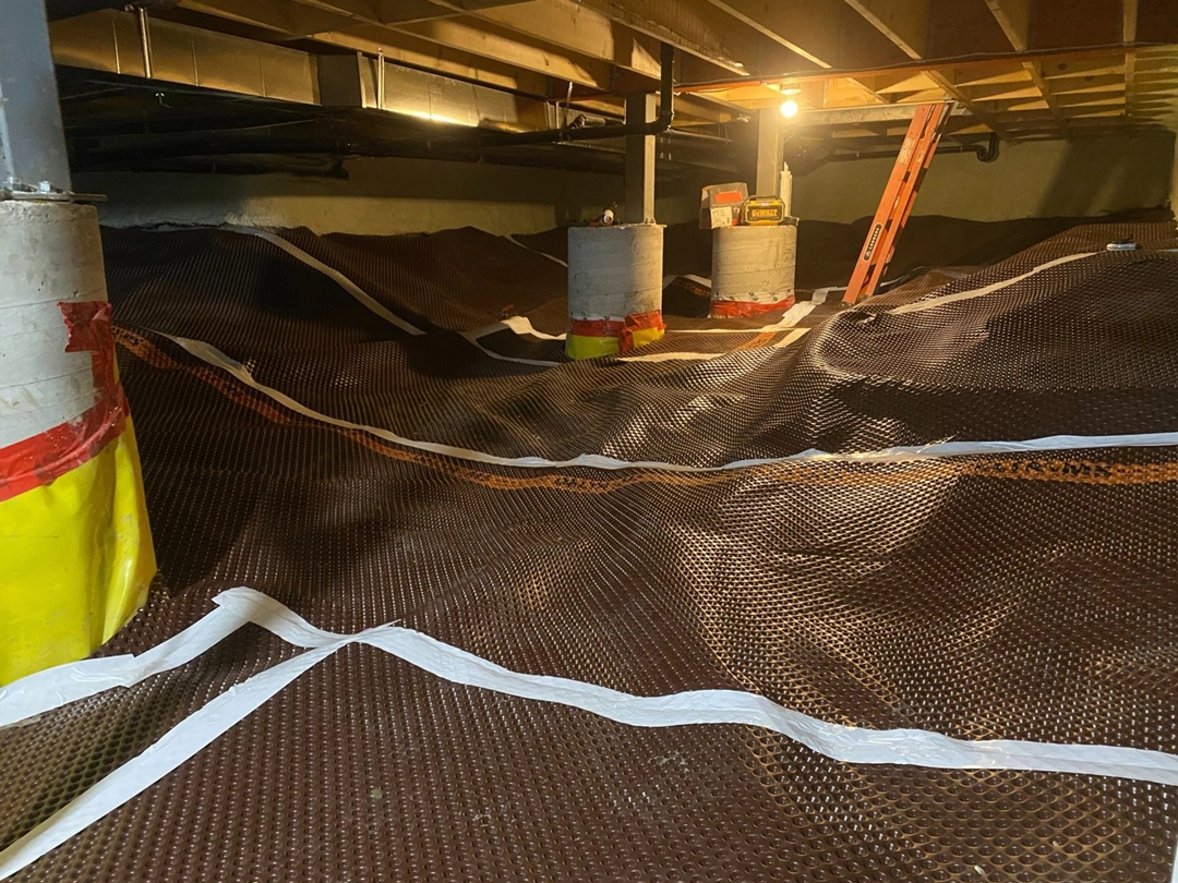 Underground space with a brown tarp covering uneven ground, yellow and red wrapped posts, and a ladder illuminated by overhead lighting