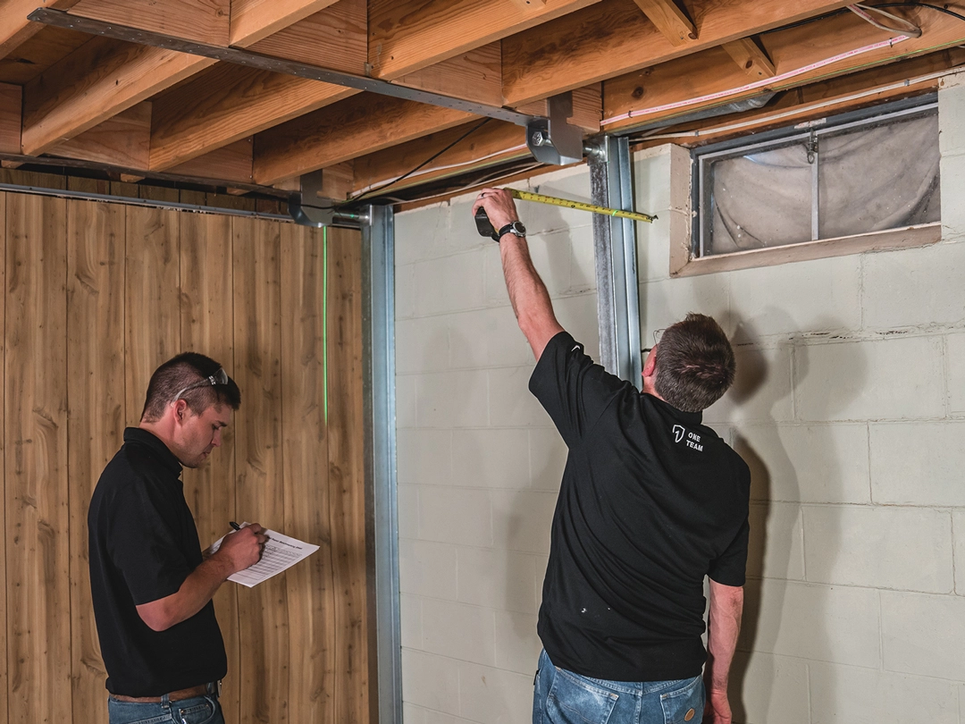 Two men measuring dimensions of a wall and ceiling.